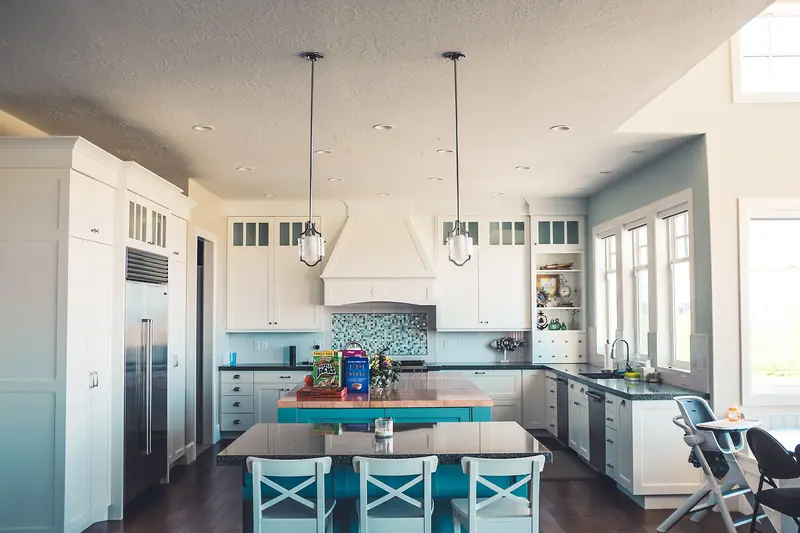 A bright white kitchen with a large island, blue bar stools, glass pendant lights and stainless appliances