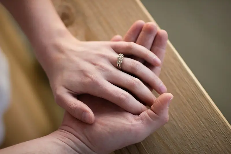A groom gently holds his bride's hand, showing her engagement ring, on a wooden table